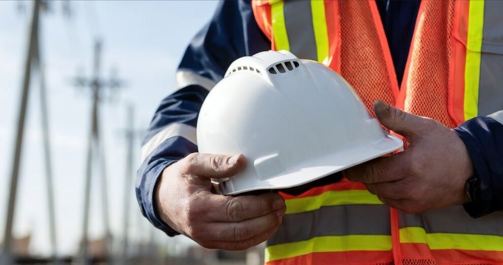 A worker in a safety vest holds a white hard hat.