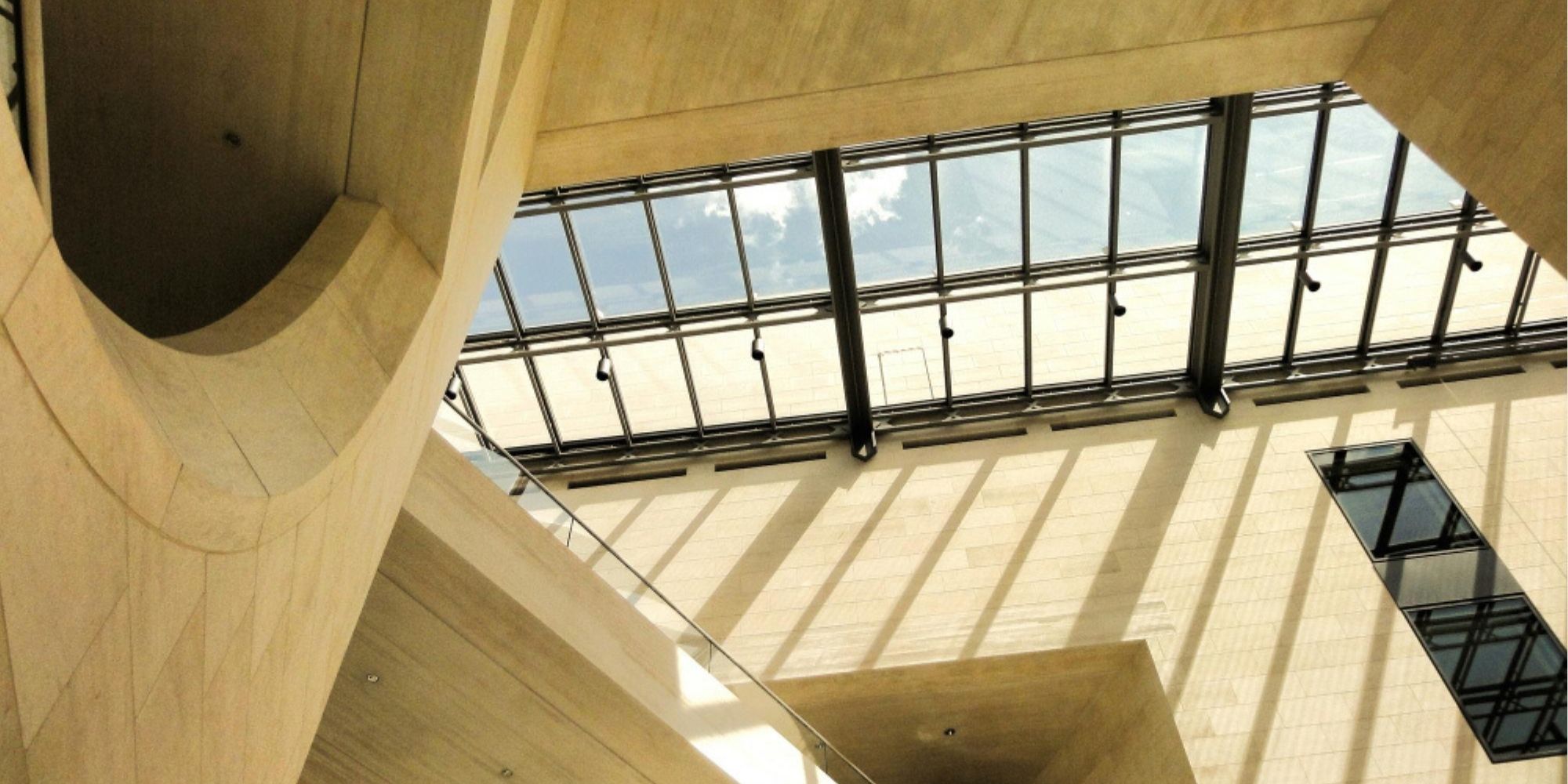 Modern building interior with large glass skylight, natural sunlight casting geometric shadows on cream-colored walls and floors, and contemporary architectural design.