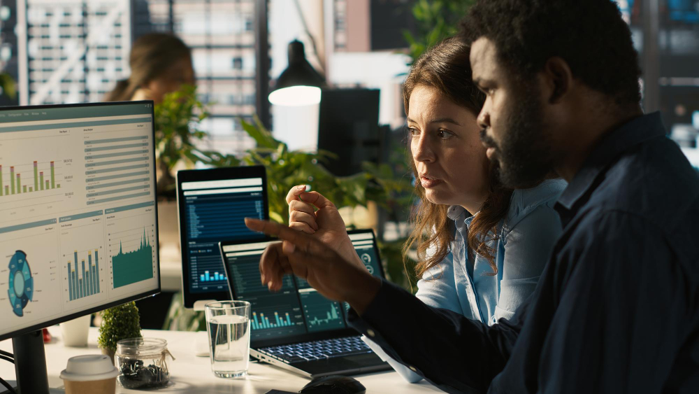 Two professionals analyzing charts, dashboards, and business data on computer screens in a modern office.