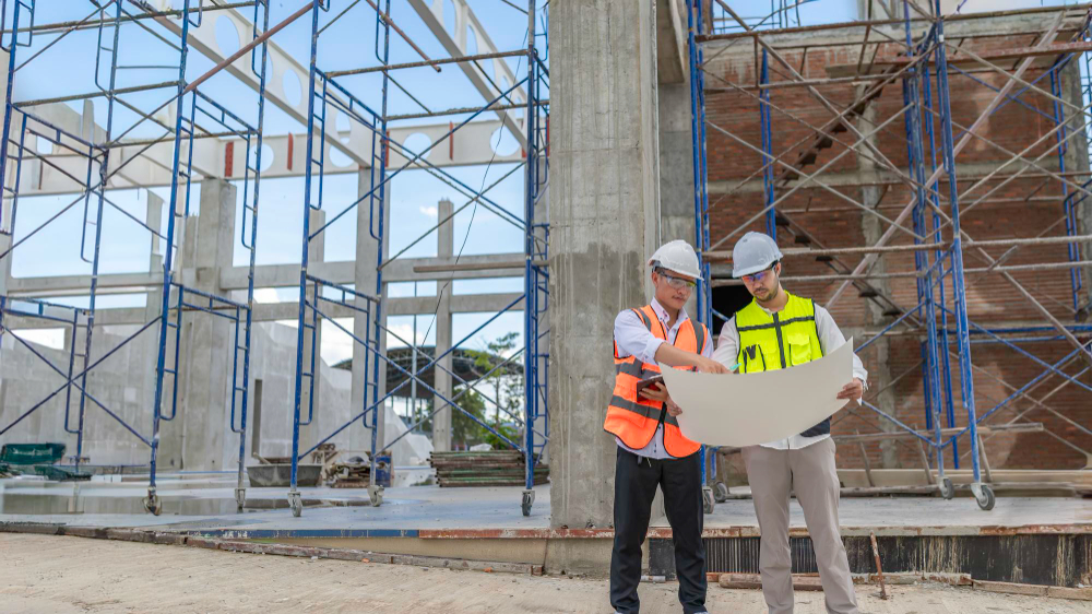 Two construction engineers wearing hard hats and safety vests reviewing blueprints at a building construction site.